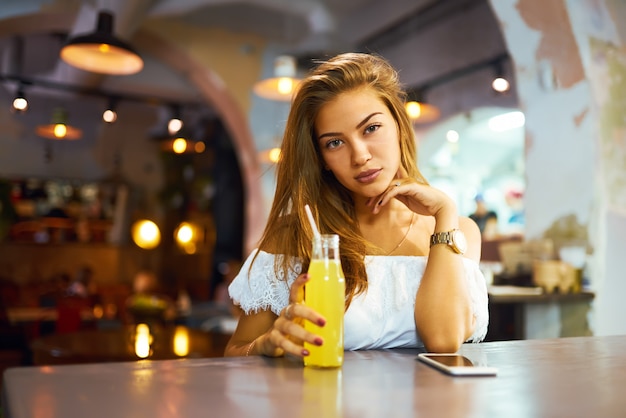 Giovane, bella ragazza con un bel sorriso seduto in un bar a bere limonata e usando un telefono