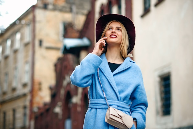 Giovane bella ragazza con un sorriso perfetto in un cappotto blu e cappello bordeaux che cammina per strada e parla al telefono cellulare