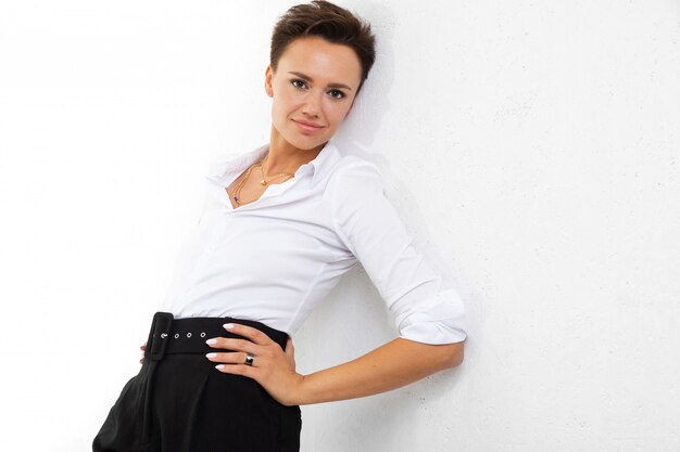 A young beautiful girl with short dark hair, makeup in a white shirt, black pants, with a wristwatch, bracelet and white manicure stands near a white wall and smiles