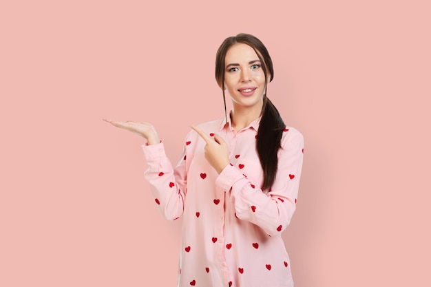 Young beautiful girl with a smile on a pink background in a pink shirt with red hearts points his fingers to the place for promotions offers or advertising