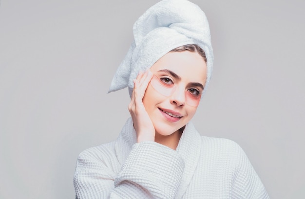 Young beautiful girl with a towel on head and a white robe girl cares for the skin using patches under her eyes Brunette spa girl Eyes mask cosmetic patches woman face closeup