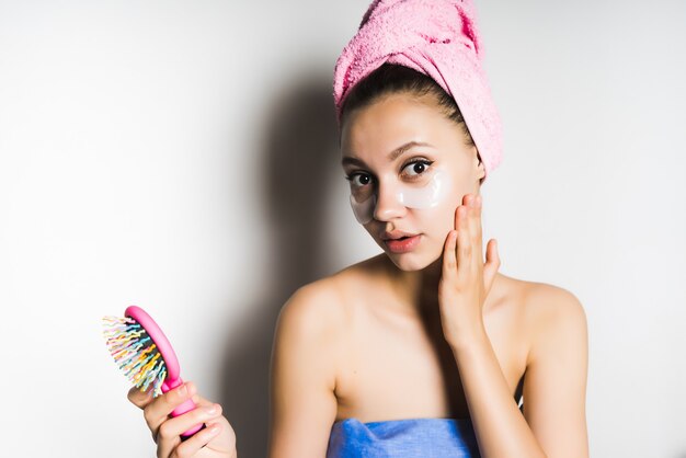 Young beautiful girl with a towel on her head looks surprised at the camera after a bath, holds in her hand a pink comb