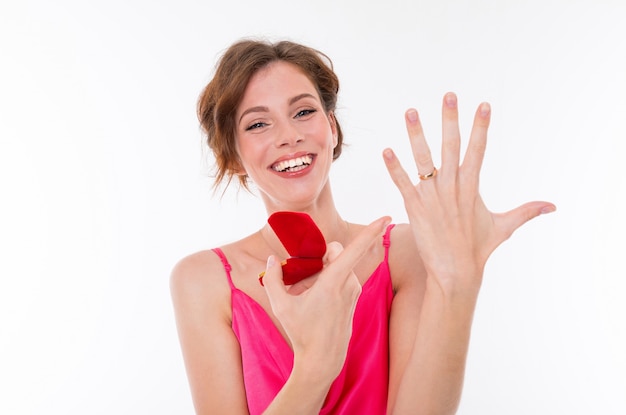 A young beautiful girl with wavy brown patched hair, clean skin, flat teeth, a beautiful smile, in a pink jersey, holds a training ring box, shows off the engagement ring on her hand and rejoices