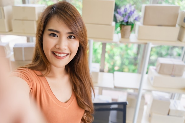 Young beautiful happy asian business woman in casual wear with smiley face is selfie at her startup home office with parcel box on shelf, SME, shopping online 