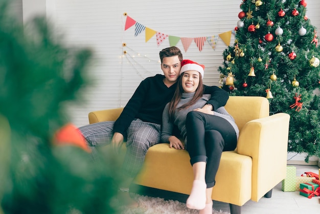 Young beautiful happy Asian woman wearing a Santa Claus hat sitting together with her boyfriend on the sofa at home with a Christmas tree in the background Image with copy space