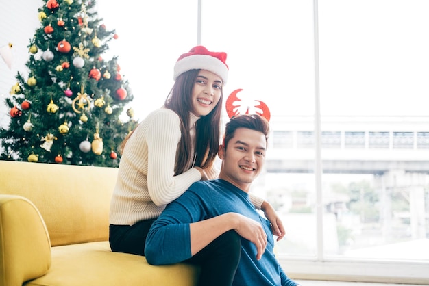 Young beautiful happy Asian woman wearing a Santa Claus hat smile at the back of her boyfriend and looks a the camera at home with a Christmas tree in the background Image with copy space