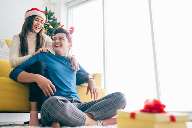 Young beautiful happy asian woman wearing santa claus hat\
surprises her boyfriend with a christmas gift at home with\
christmas tree in the background image with copy space