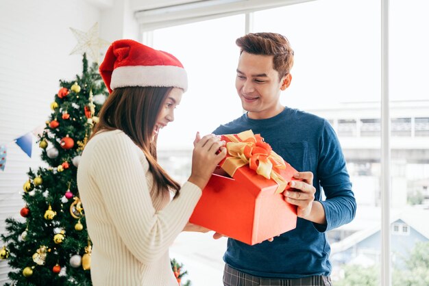 Young beautiful happy Asian woman wearing Santa Claus hat surprises her boyfriend with a Christmas gift at home with Christmas tree in the background Image with copy space