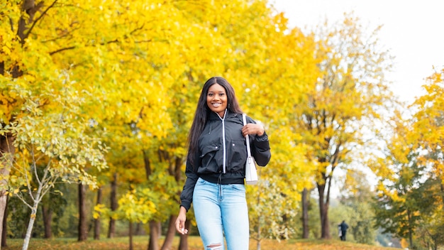 Giovane bella ragazza nera felice in abiti casual alla moda guarda con borsetta alla moda passeggiate nel parco con fogliame autunnale dorato brillante