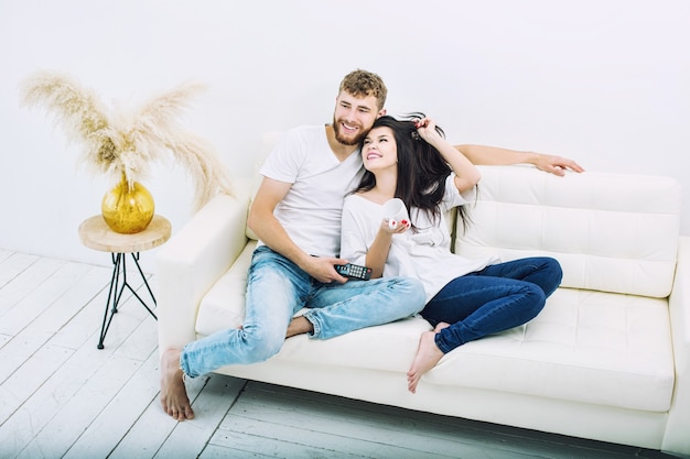 Young beautiful and happy couple man and woman at home on white sofa watching tv smiling and hugging