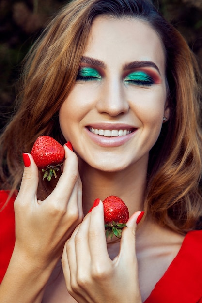 Young beautiful happy funny girl with red dress and makeup holding strawberry in summertime in the park. healthy lifestyle, diet beauty and happiness concept.