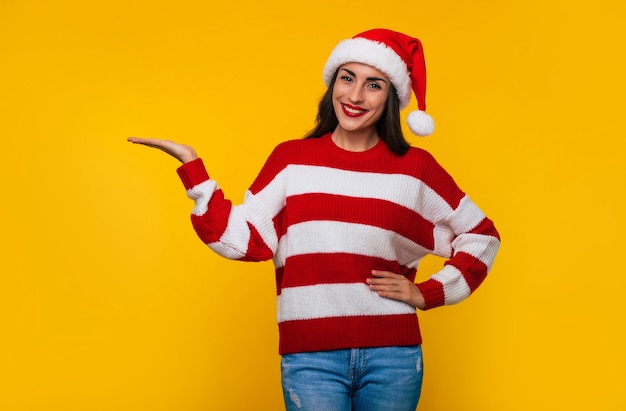 La giovane bella ragazza felice in cappello di babbo natale e maglione invernale è in posa isolata su sfondo giallo nel periodo natalizio