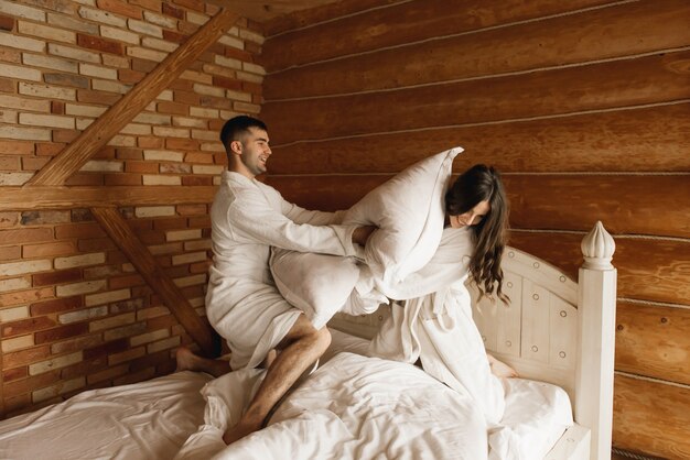 Young beautiful happy lovers newlyweds in white bath robes fighting with pillows on the bed