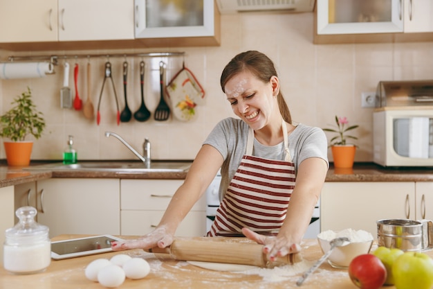 The young beautiful happy woman sitting at a table with flour and tablet, rolling a dough with a rolling pin and going to prepare a cakes in the kitchen. Cooking home. Prepare food.