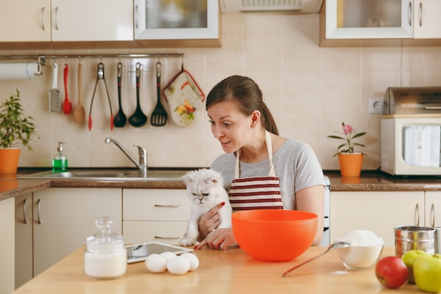 The young beautiful happy woman with a white Persian cat looking for a recipe of cakes in a tablet in the kitchen. Cooking home. Prepare food.