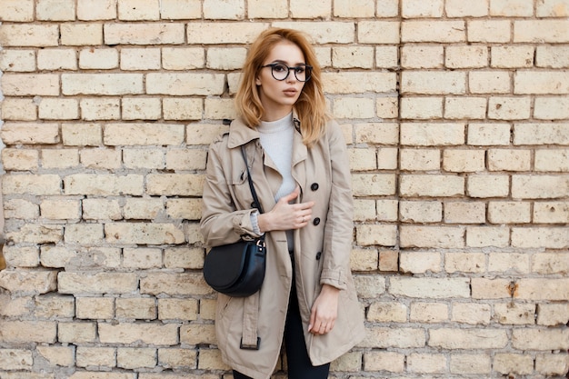 Young beautiful hipster woman in stylish glasses in a trendy spring coat in a white T-shirt with a fashionable black leather bag stands near the vintage brick wall. Charming sweet girl.