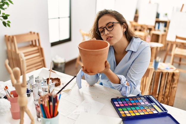 Young beautiful hispanic woman artist painting ceramic pot at art studio