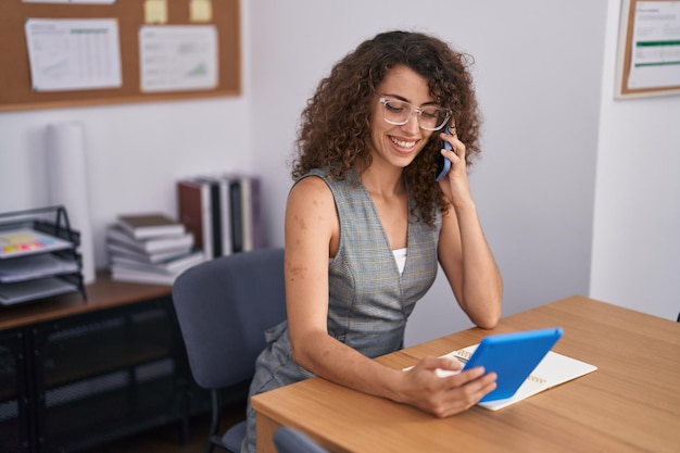 Young beautiful hispanic woman business worker talking on smartphone using touchpad at office