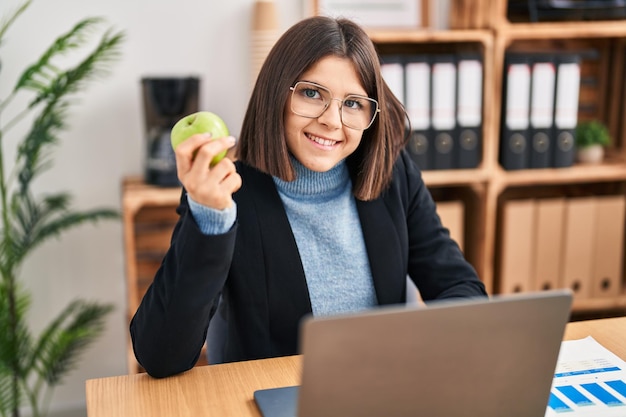 Young beautiful hispanic woman business worker using laptop eating apple at office