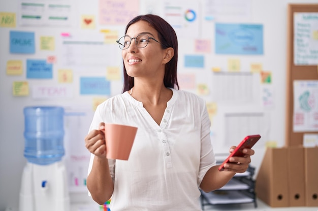 Young beautiful hispanic woman business worker using smartphone drinking coffee at office