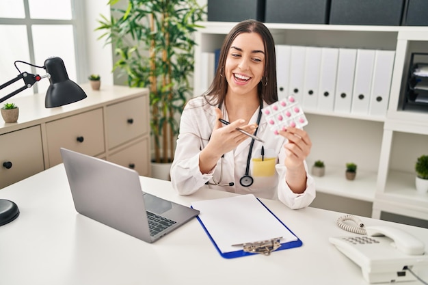 Young beautiful hispanic woman doctor using laptop holding pills at clinic