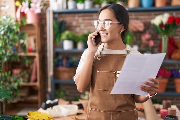 Young beautiful hispanic woman florist talking on smartphone reading document at florist