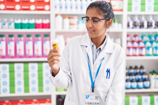 Young beautiful hispanic woman pharmacist smiling confident holding pills bottle at pharmacy