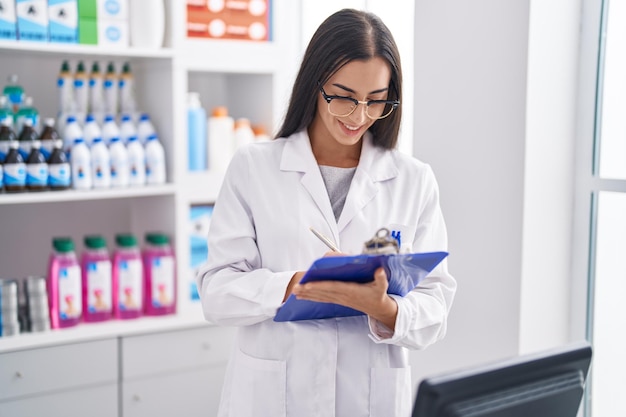 Young beautiful hispanic woman pharmacist writing on document at pharmacy