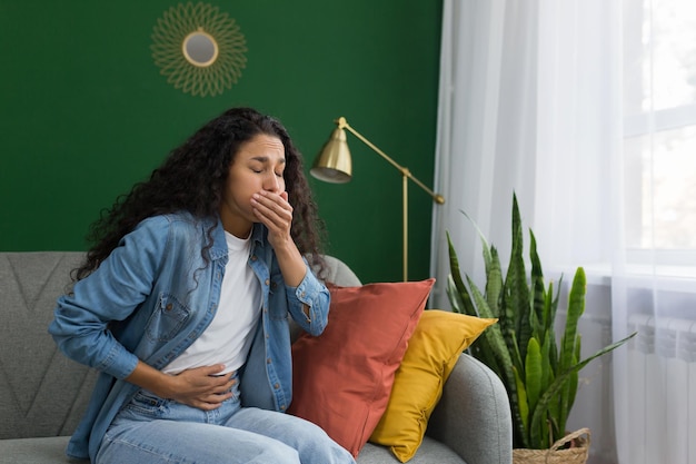 Young beautiful hispanic woman sick at home sitting on sofa with nausea and stomach ache sitting on