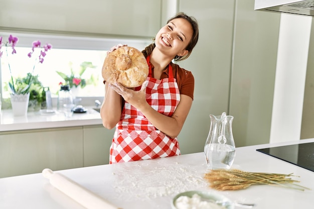 Photo young beautiful hispanic woman smiling confident holding bread at the kitchen