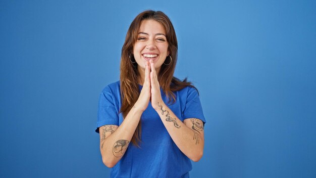 Young beautiful hispanic woman smiling confident praying with hands together over isolated blue background