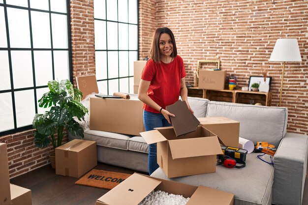 Young beautiful hispanic woman smiling confident unpacking cardboard box at new home