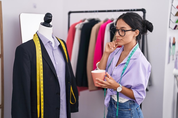 Young beautiful hispanic woman tailor drinking coffee measuring jacket at tailor shop