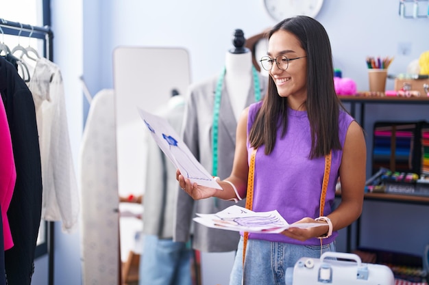 Young beautiful hispanic woman tailor smiling confident looking clothing design at tailor shop