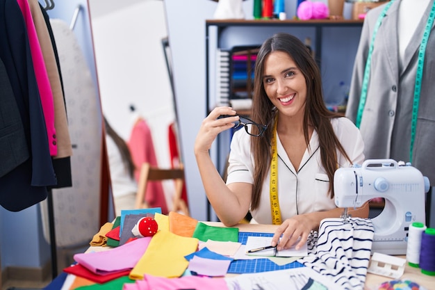 Young beautiful hispanic woman tailor smiling confident sitting on table at clothing factory