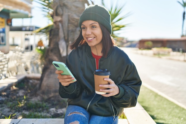 Young beautiful hispanic woman using smartphone drinking coffee at park