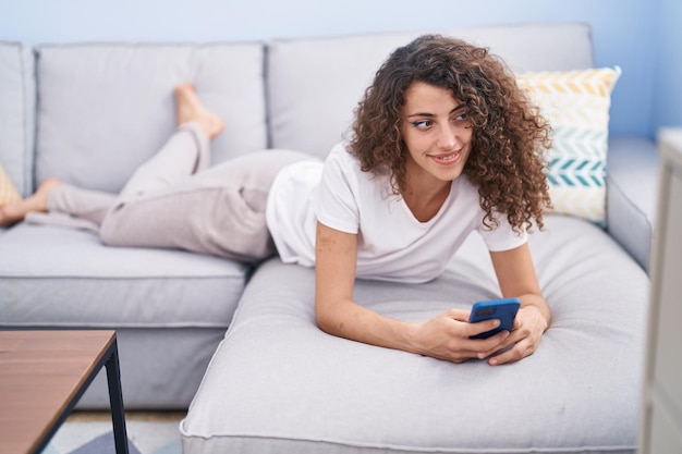 Young beautiful hispanic woman using smartphone lying on sofa at home