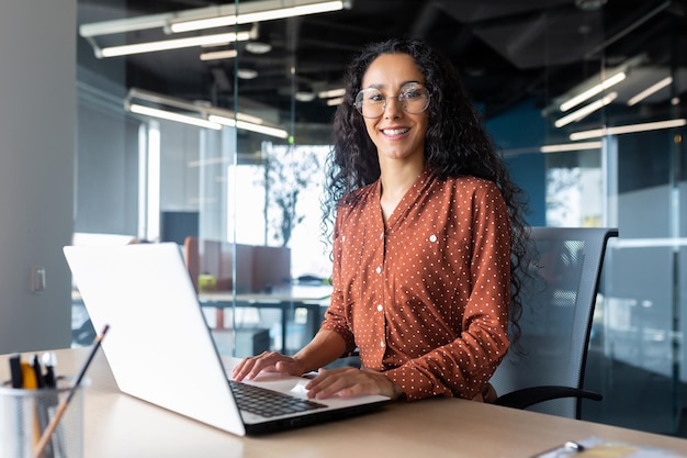 Young beautiful hispanic woman working inside modern office businesswoman smiling and looking at