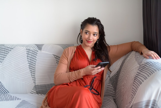 Young beautiful Indian woman thinking while using phone in the living room at home