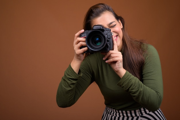 Young beautiful Indian woman with camera against brown wall