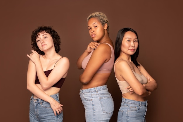 Young beautiful intercultural females in tanktops and blue jeans standing in row on brown