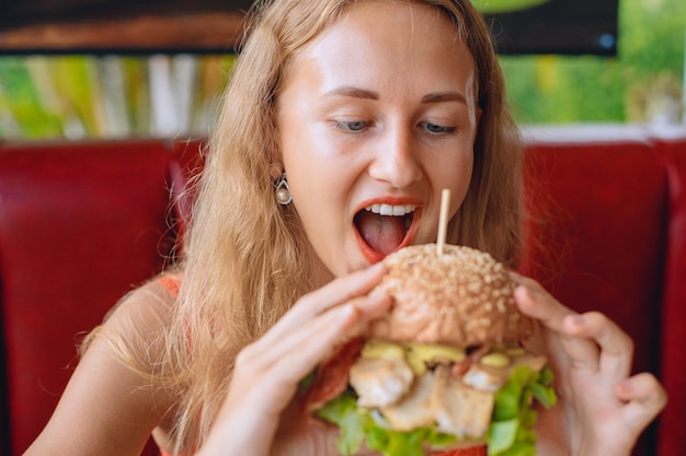 ブロンドの髪の若い美しい女性は、カフェで巨大なハンバーガーを噛んでいます。ファーストフードのコンセプト。ポートレート。