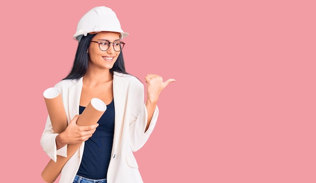 Young beautiful latin girl wearing architect hardhat holding blueprints pointing thumb up to the side smiling happy with open mouth