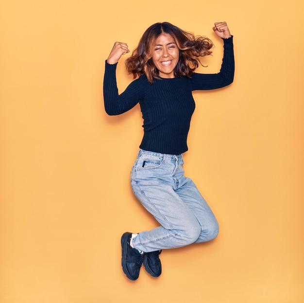 Young beautiful latin woman smiling happy. Jumping with smile on face celebrating with fists up over isolated yellow background