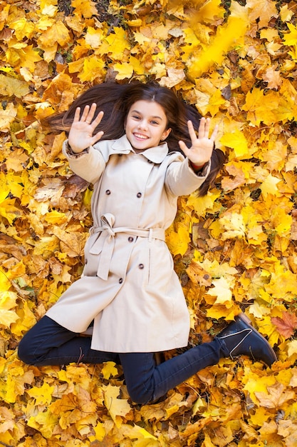 Фото Молодая красивая девочка в бежевом пальто лежит на желтых листьях вид сверху в осеннем парке