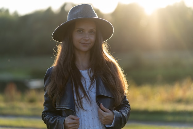 A young, beautiful long-haired woman in a hat smiles, looks at the camera in the park in the bright glow of sunset rays