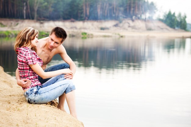 Young beautiful loving couple having rest on river shore on summer day with green nature at background