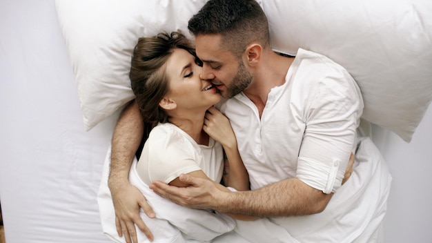 Young beautiful and loving couple kiss and hug into bed while waking up in the morning