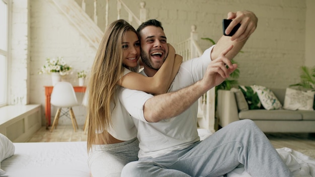 Young beautiful and loving couple take selfie picture on smartphone camera while sitting in bed