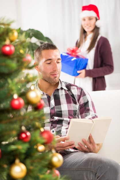 Young beautiful man reading book while his girlfriend holds a Christmas gift and she wants to surprise him.